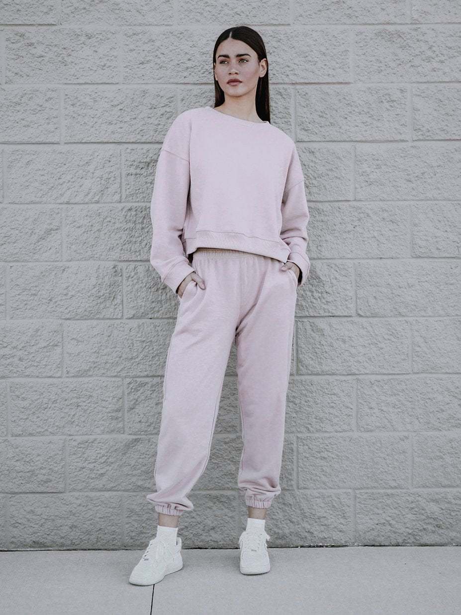 A person stands against a light gray brick wall, wearing a trendy boxy silhouette pink sweatshirt and matching sweatpants set, crafted from a luxe cotton blend, paired with white sneakers. Their hands are in their pockets and they are looking slightly to the left.