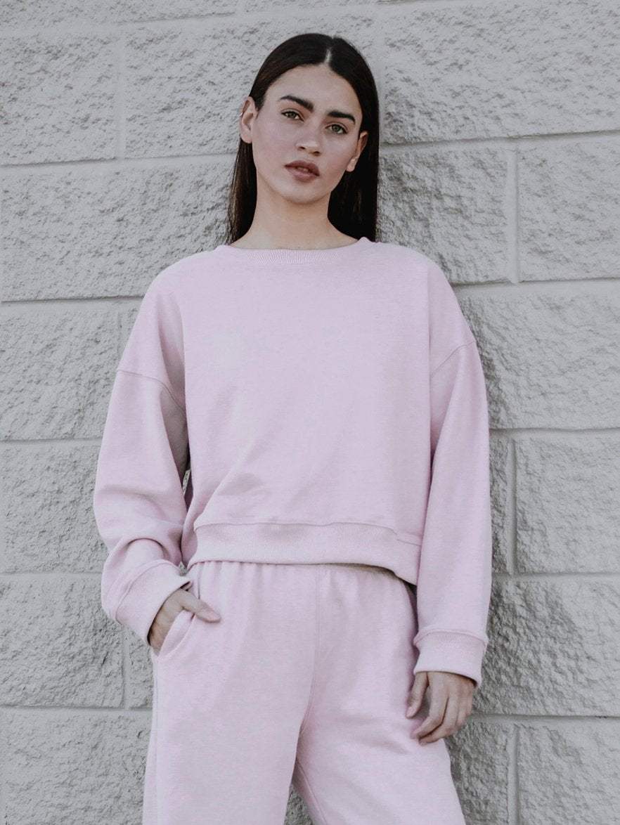 A person with long hair stands against a textured brick wall, wearing a matching light pink sweatshirt and sweatpants made from a luxe cotton blend. They have their hands in their pockets, staring confidently at the camera. Completing the look, they sport stylish white shoes.