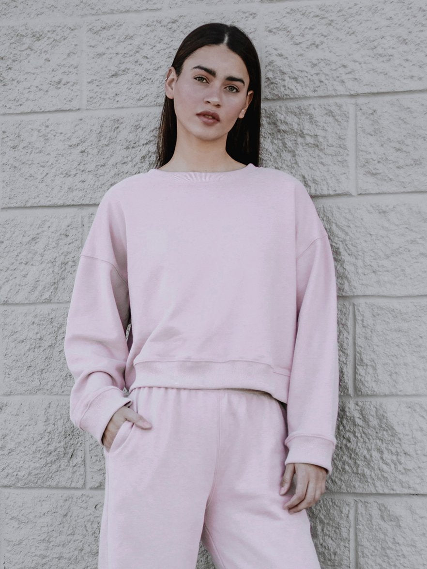 A person is standing against a textured brick wall, wearing a light pink boxy silhouette sweatshirt and matching joggers.