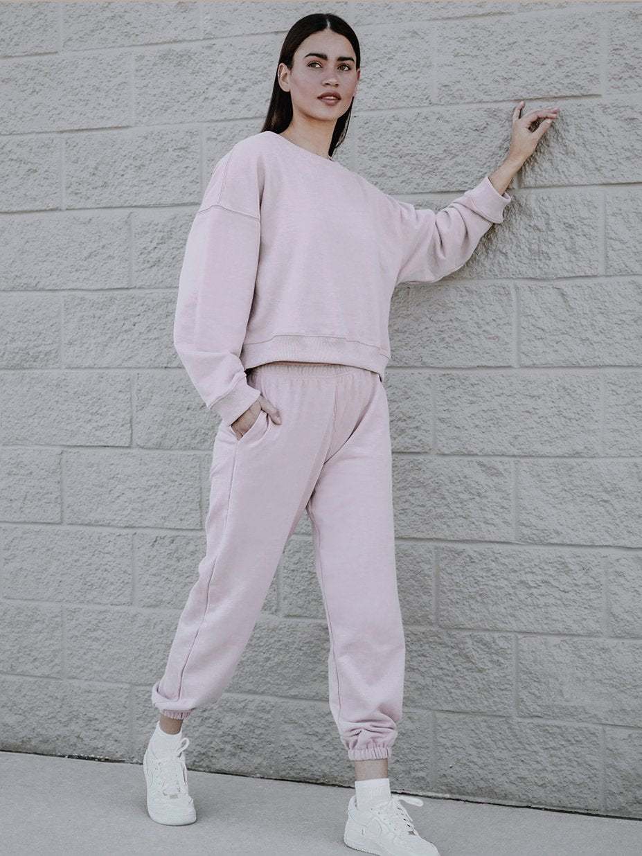 A person is standing against a textured brick wall, wearing a light pink boxy silhouette sweatshirt and matching joggers. They have one hand resting on the wall and are looking forward. They are also wearing white sneakers.