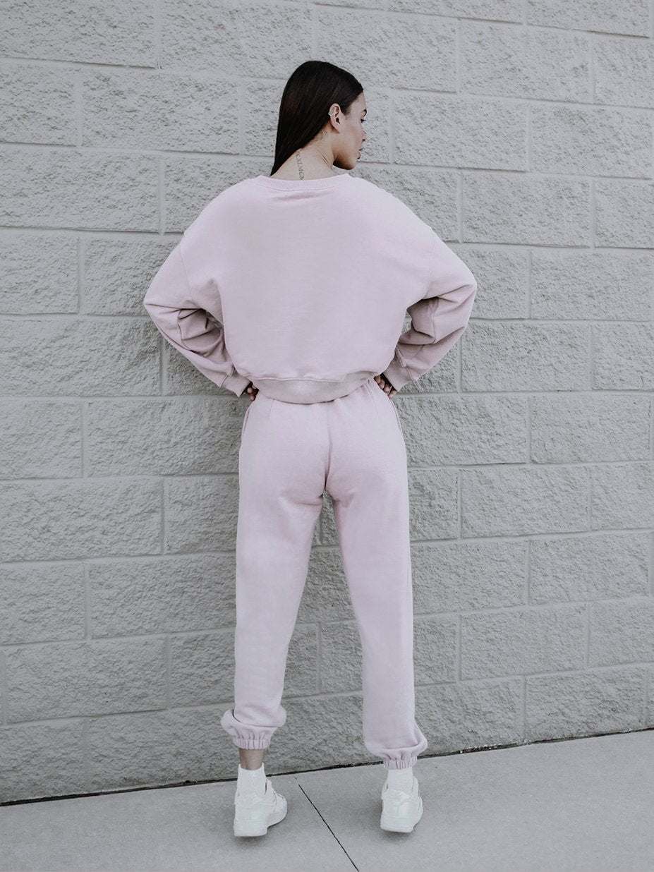A person stands with their back to the camera in front of a textured, light gray wall. They are wearing a light pink, long-sleeve sweatshirt and matching sweatpants featuring a boxy silhouette, paired with white sneakers. Their hands rest on their hips.