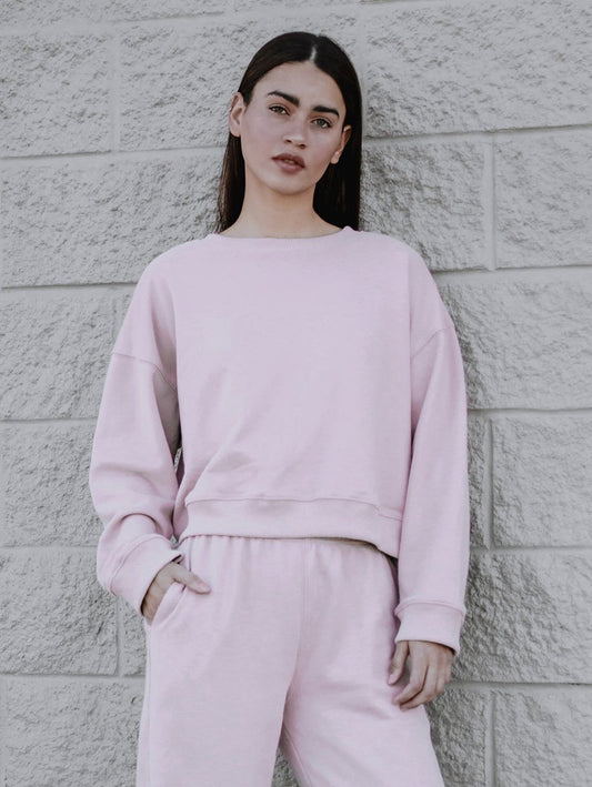 A person is standing against a textured brick wall, wearing a light pink boxy silhouette sweatshirt and matching joggers. 